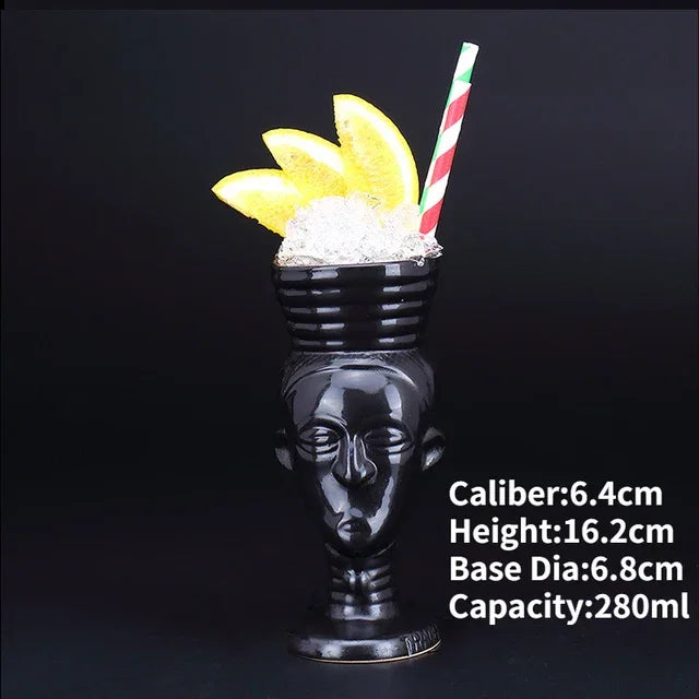 Unique black ceramic face-shaped mug with crushed ice, orange slices, and a striped straw, perfect for stylish décor or creative cocktails.